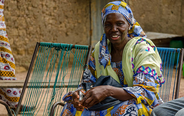 Bamako, Mali – August 18, 2013: An African woman smiling whilst sitting on a chair in front of her house with her typical African clothing and her phone in her hand.